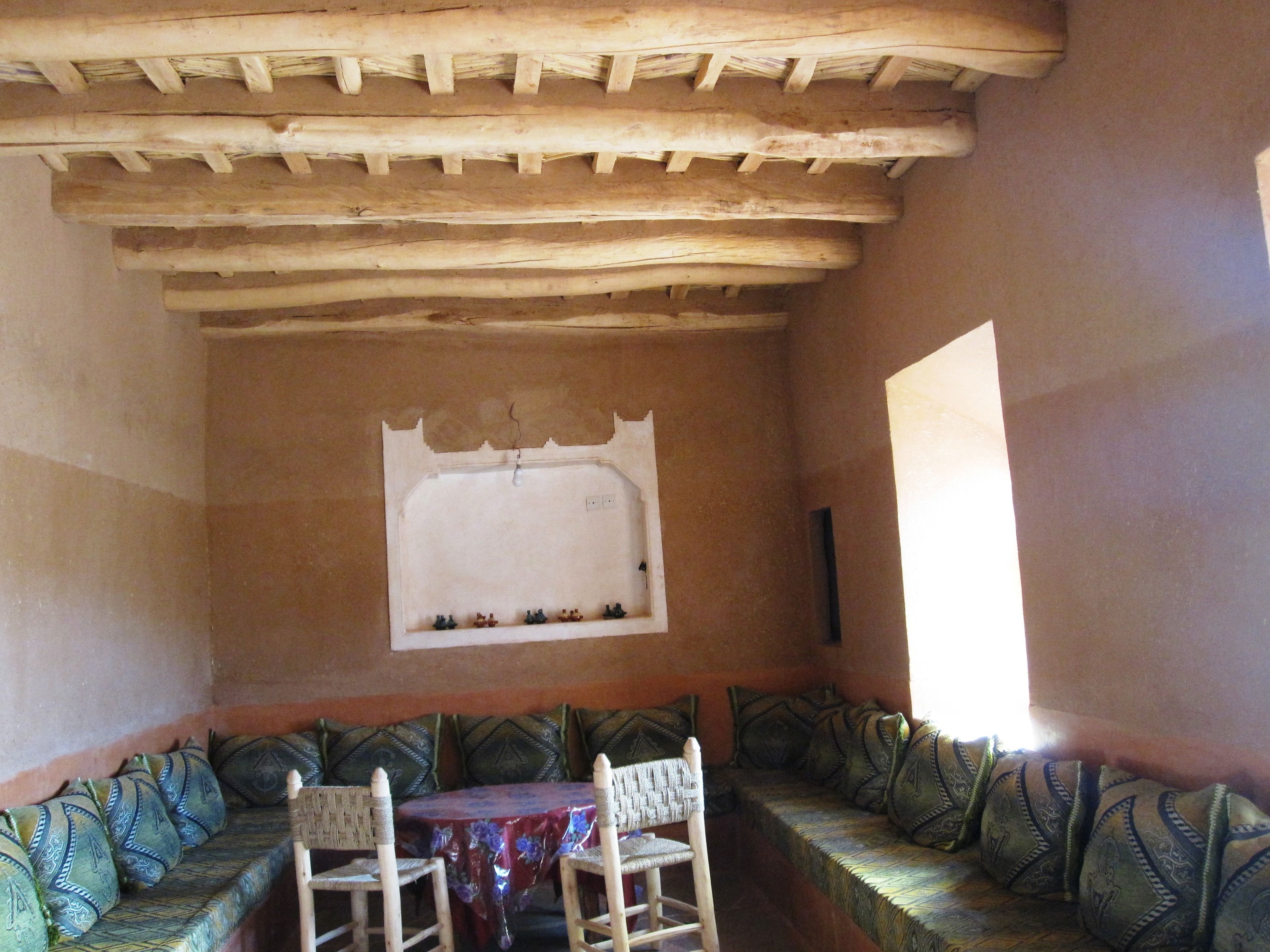 Quarto com teto de madeira, janela com moldura decorativa, mesa com cadeiras e sofá com almofadas estampadas.