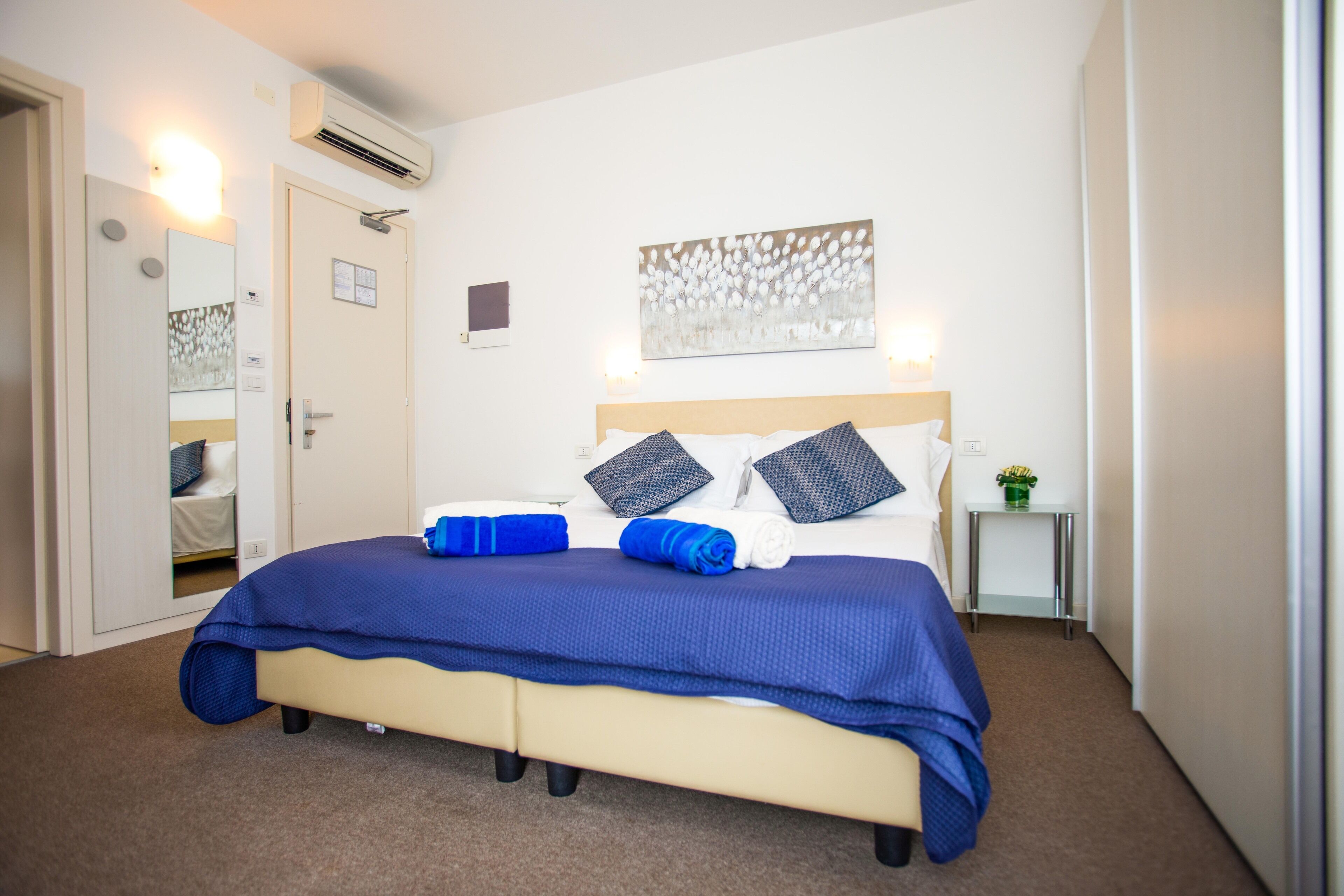 A neatly made bed with a blue bedspread and pillows, a bedside table with a lamp, and a wall-mounted air conditioner.