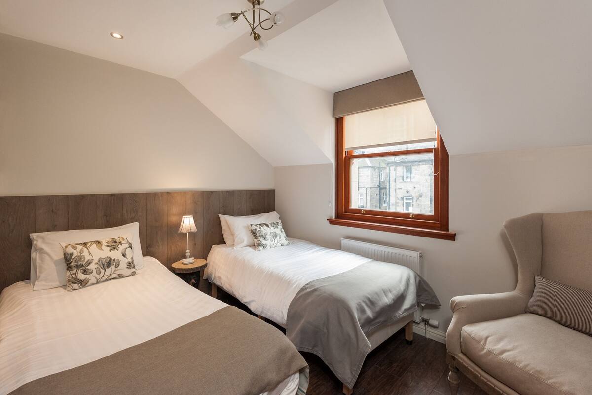 A bedroom with two beds, a nightstand, a lamp, a chair, and a window with a view of buildings.