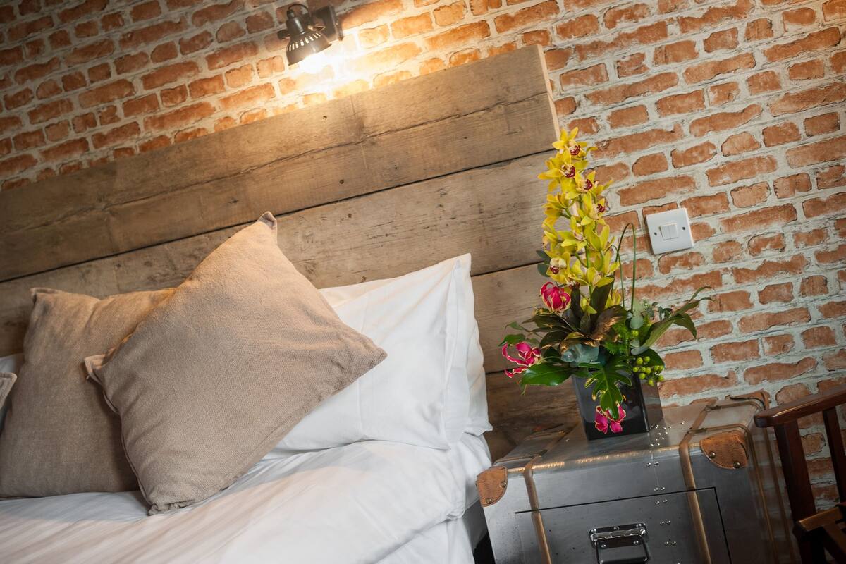 A bed with white bedding and beige pillows, a wooden headboard, a small table with a vase of flowers, and a brick wall.