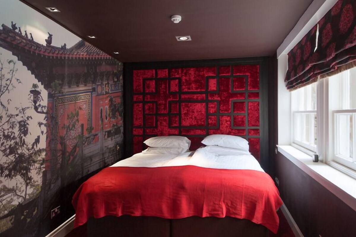 A bedroom with a red bedspread, a mural on the wall, and a window with curtains.
