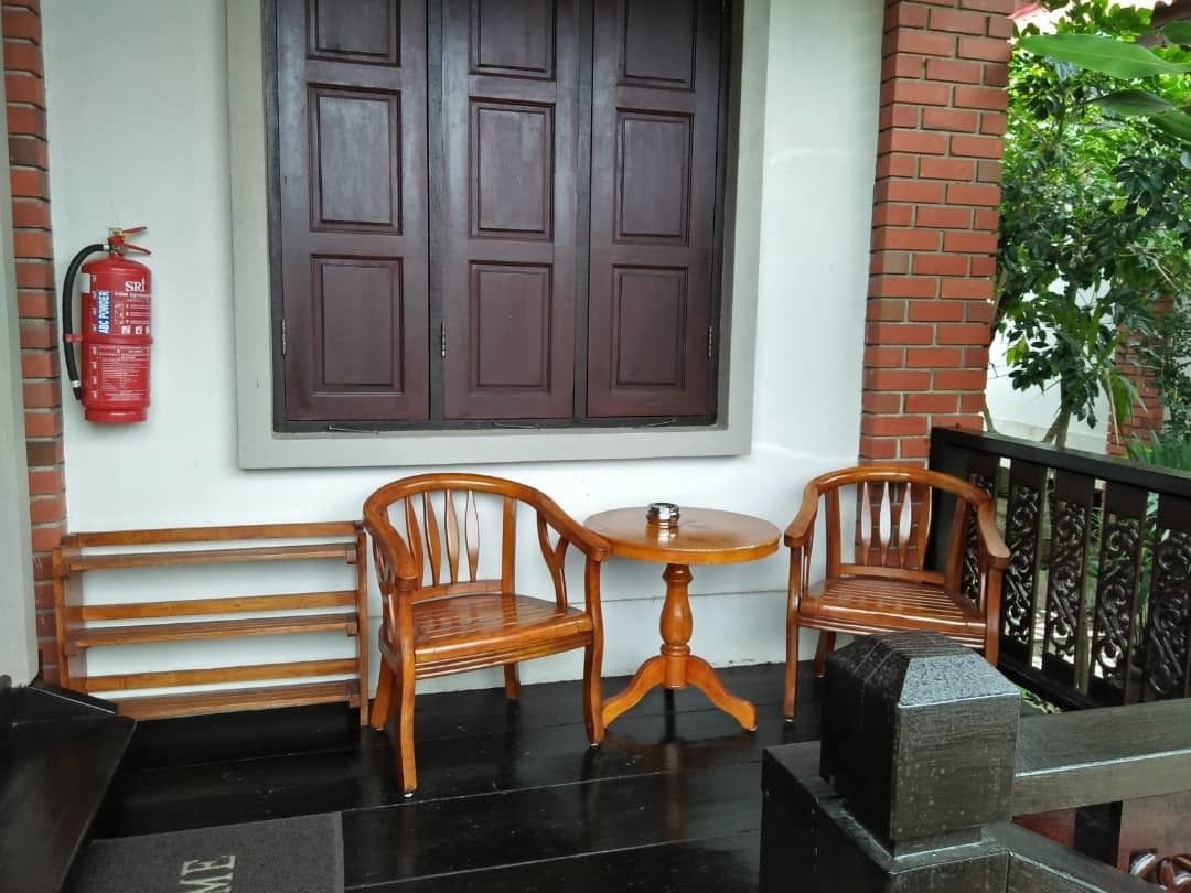 A wooden bench and two chairs with a small round table in front, a fire extinguisher on the wall, and a large window with dark wooden shutters.