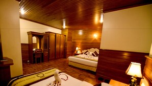 Habitación de hotel con techo de madera, cama con cubrecama blanco y pétalos de rosa rojos, mesita de noche con lámpara y armario de madera.