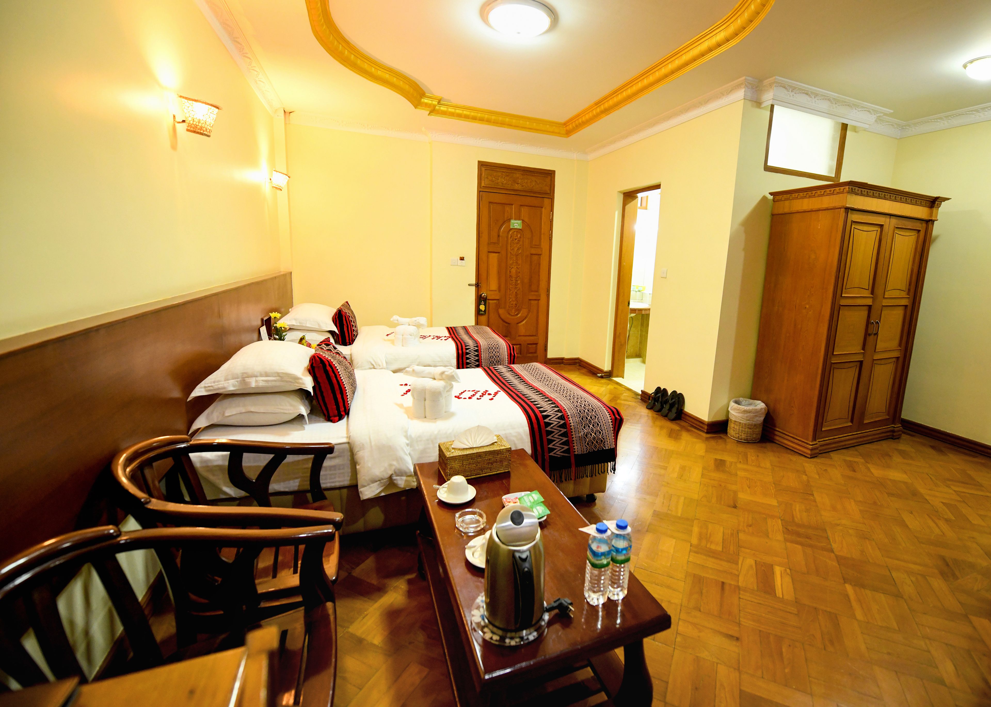A hotel room with two beds, a wooden wardrobe, a dining table with a kettle and water bottles, and a chair.