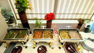A buffet with various food options, a window with vertical blinds, and decorative plants.