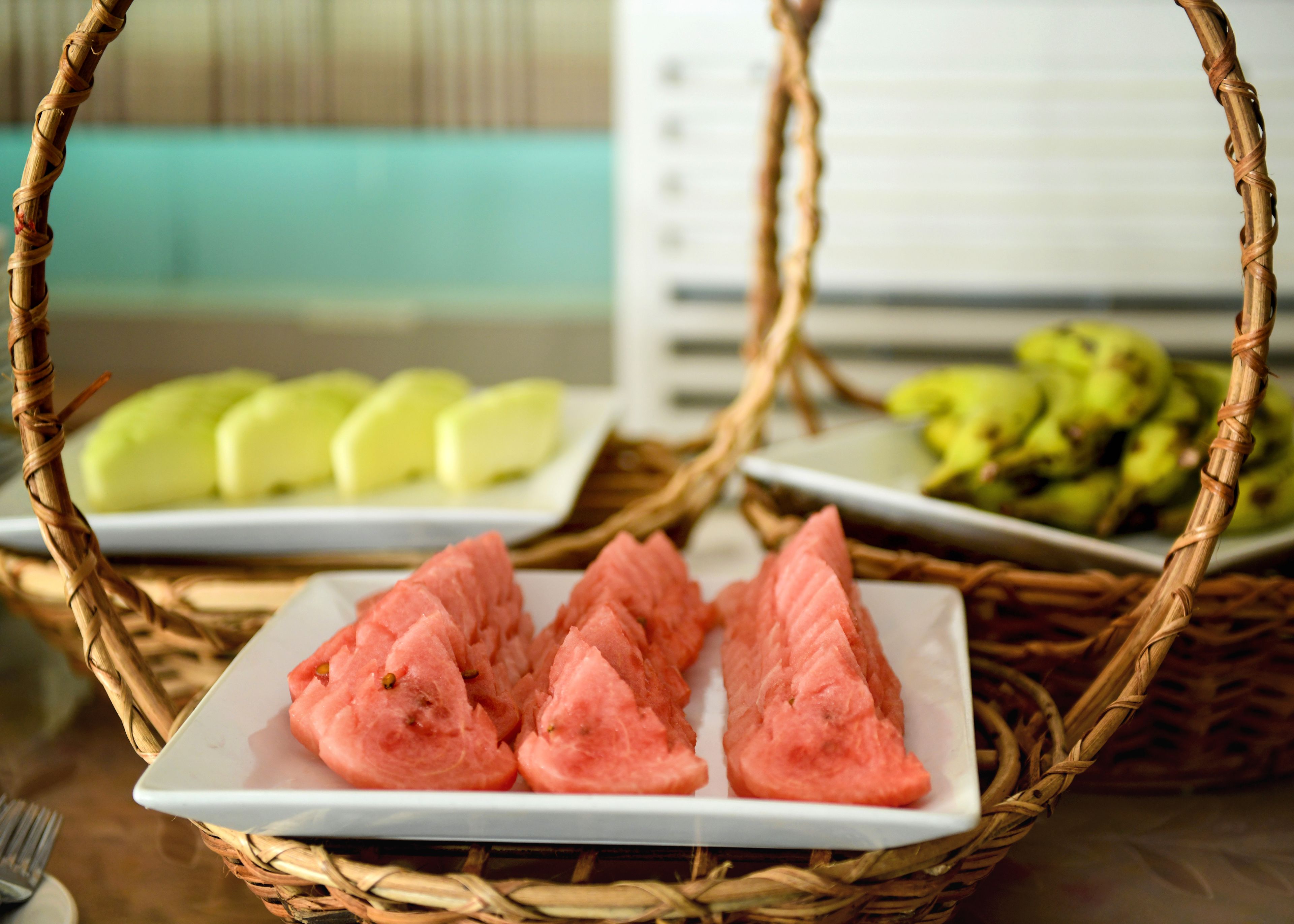 Ein Korankorb mit einem weißen Teller mit Wassermelonenstücken, einem Teller mit grünem Obst und einem Teller mit grünen Bananen.