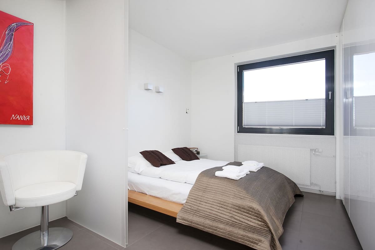 Quarto de hotel moderno com uma cama, uma cadeira branca e uma janela com persianas.