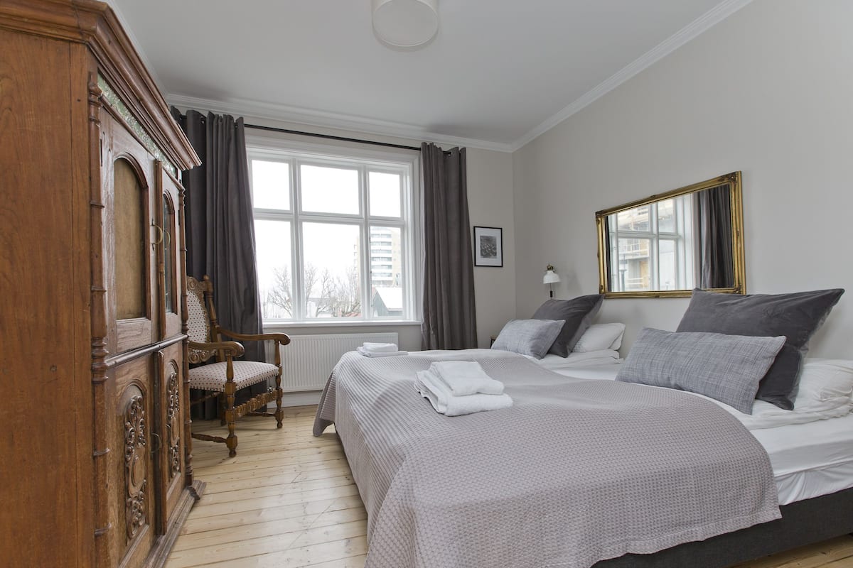 Quarto com uma cama grande, guarda-roupa de madeira, uma cadeira, janela com cortinas e espelho na parede.