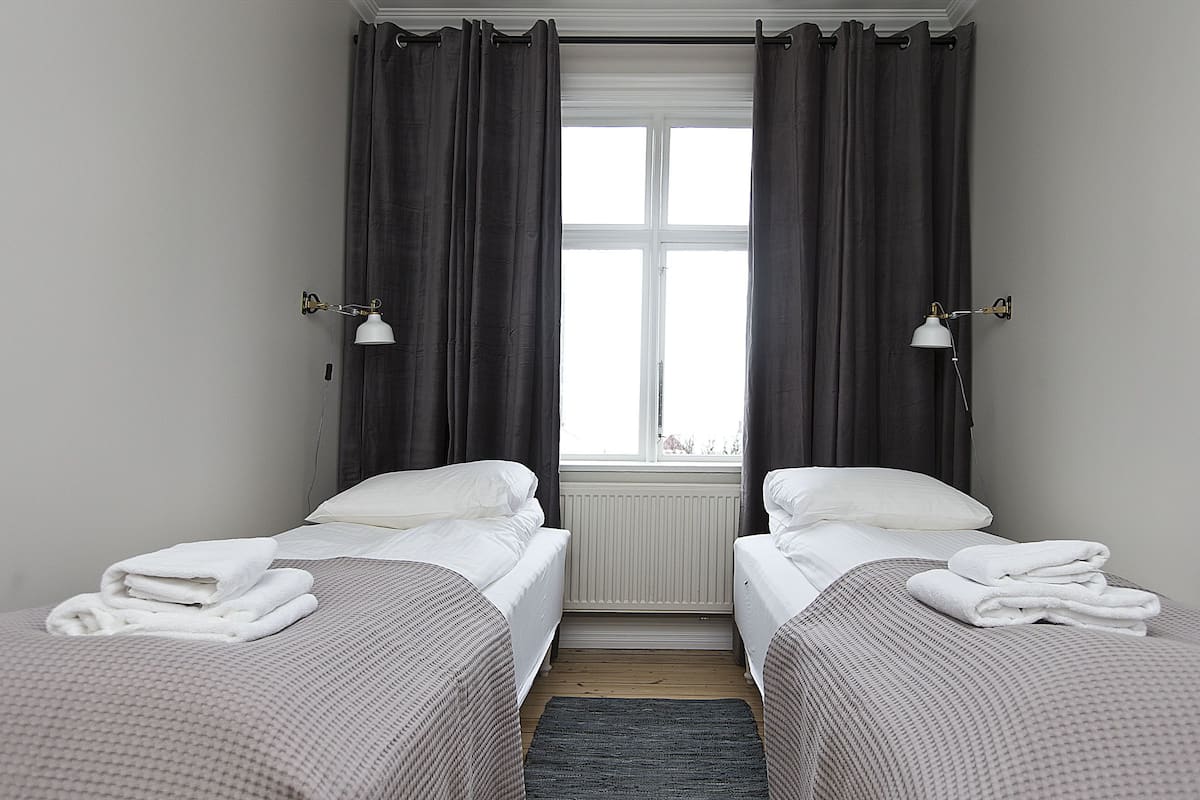Quarto de hotel com duas camas, janela com cortinas e duas luminárias fixadas na parede.