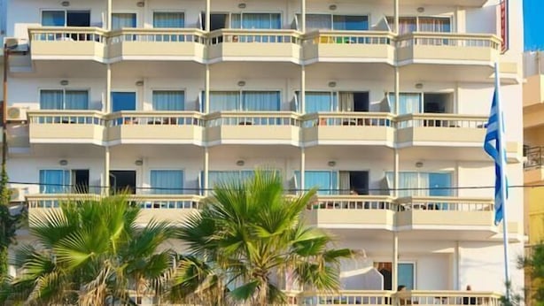 A multi-story hotel with balconies, a flag, and palm trees in the foreground. - Theo Beach Hotel (Rethymnon)