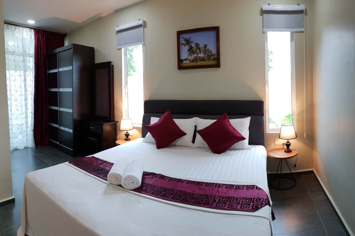 A hotel room with a large bed, two bedside lamps, a dark headboard, a wardrobe, and a window with a view of palm trees.