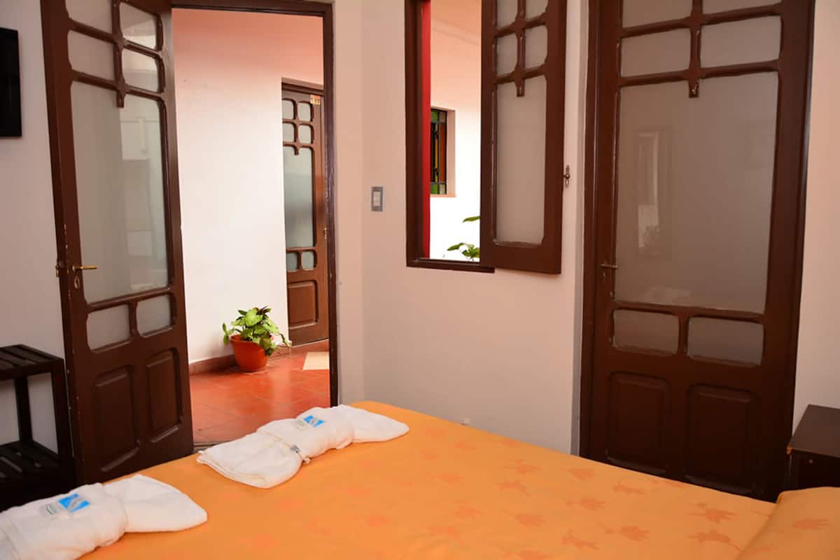 A hotel room with two open doors leading to a hallway, a bed with an orange bedspread, a potted plant, and a small table with a lamp.