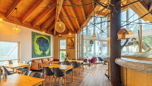 Un vaste espace dédié à la salle à manger, avec des tables et des chaises en bois, de grandes fenêtres et un plafond haut avec des poutres apparentes.