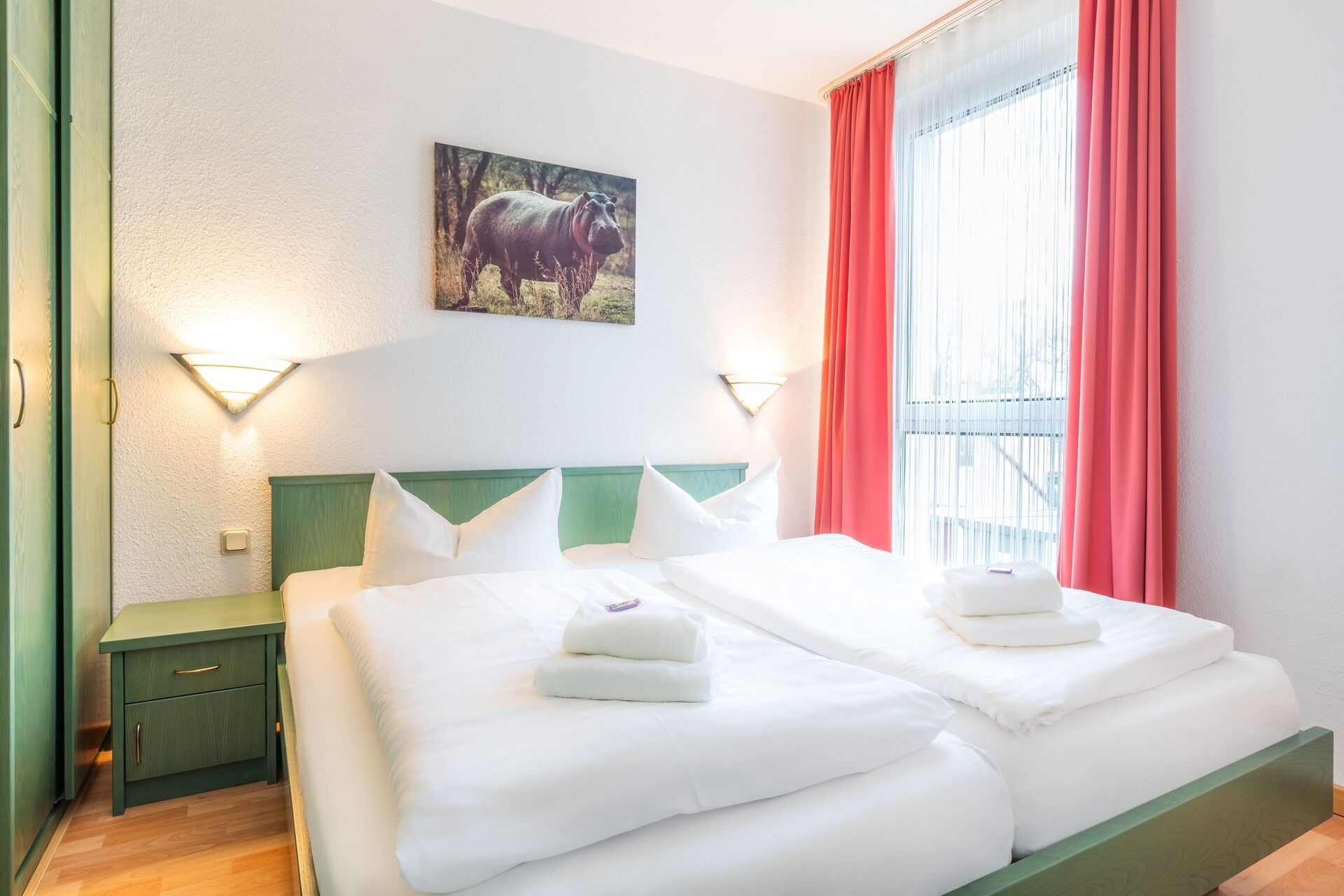 A hotel room with a double bed, two pillows, a nightstand, a wall-mounted light, a painting of a bull, and a window with red curtains.