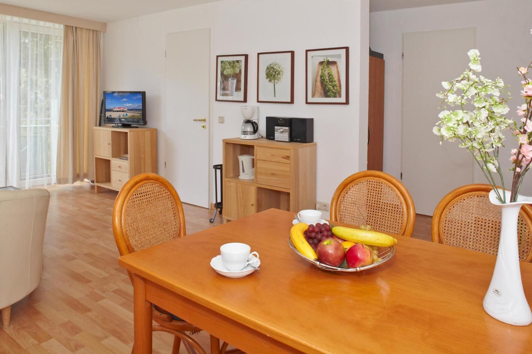 A dining area with a wooden table, four wicker chairs, a bowl of fruit, a white cup, and a vase with flowers.