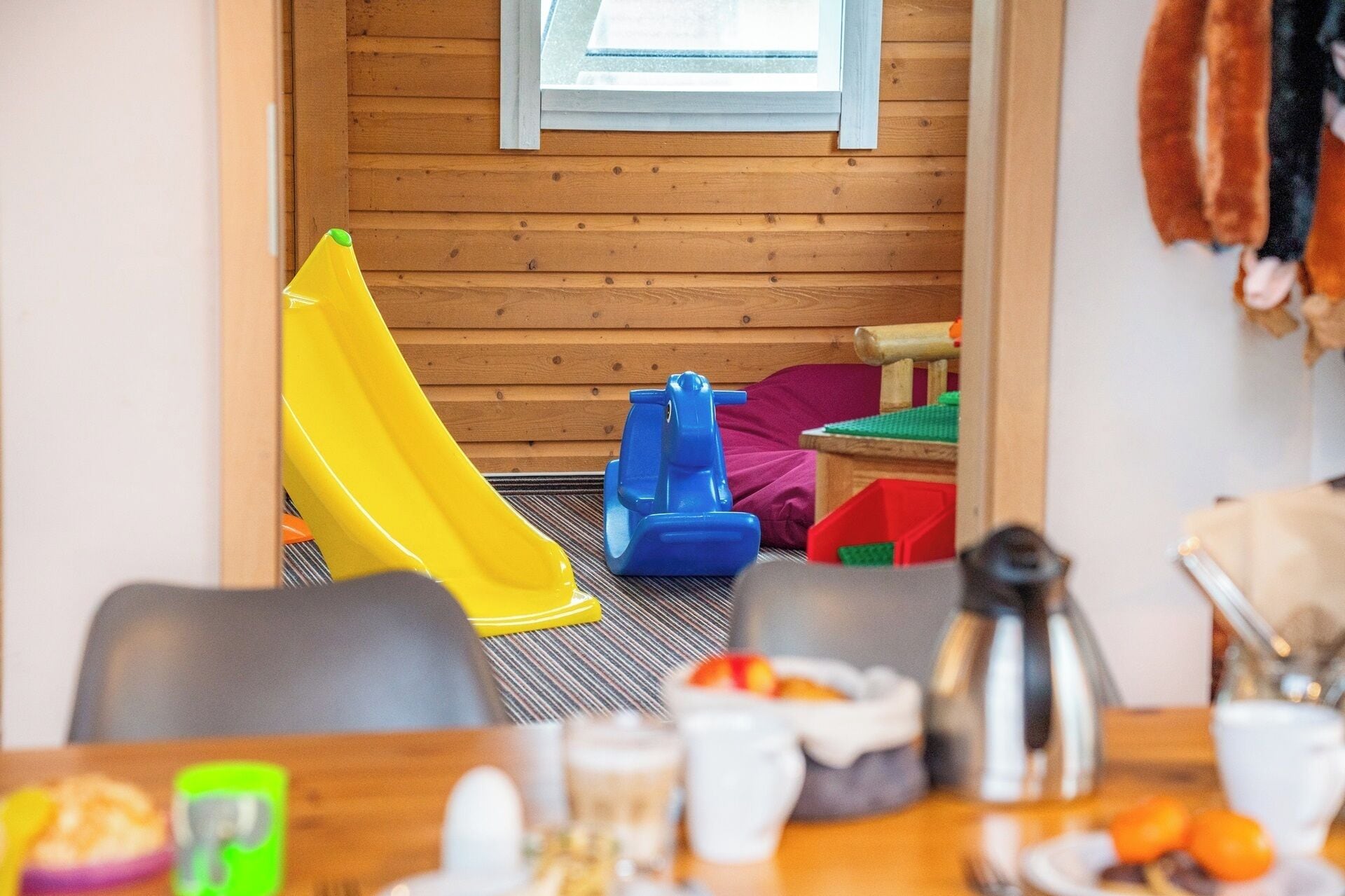 En lekplats för barn med en gul rutschkana och blå leksaksbilar, ett matbord med mat och dryck samt en vägg av trä med ett fönster.