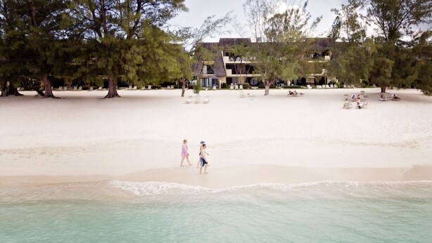 A beach with a building in the background, trees, and people walking. - The Islands Club (Seven Mile Beach)