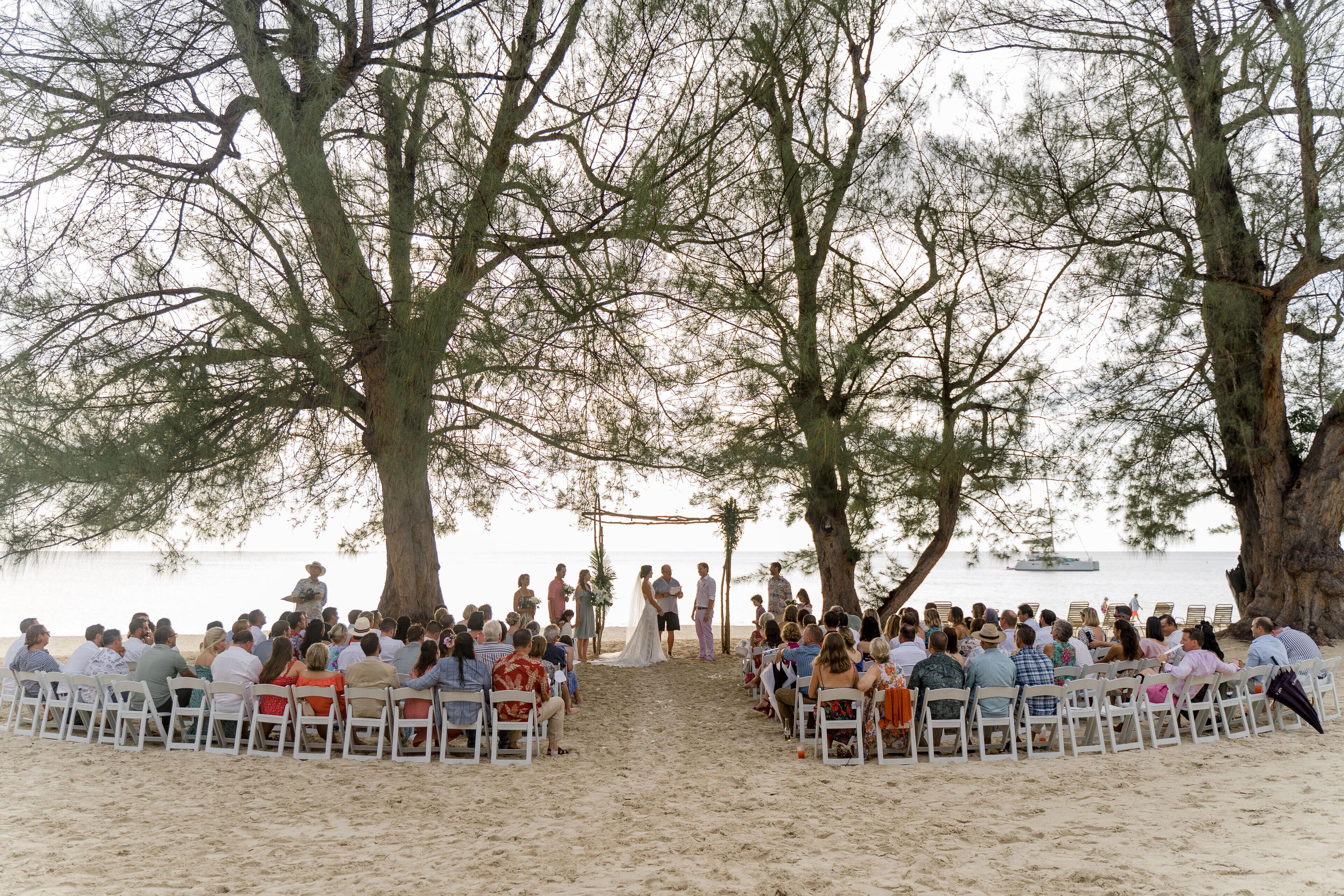 A beach wedding ceremony with guests seated on white chairs arranged in a semi-circle.