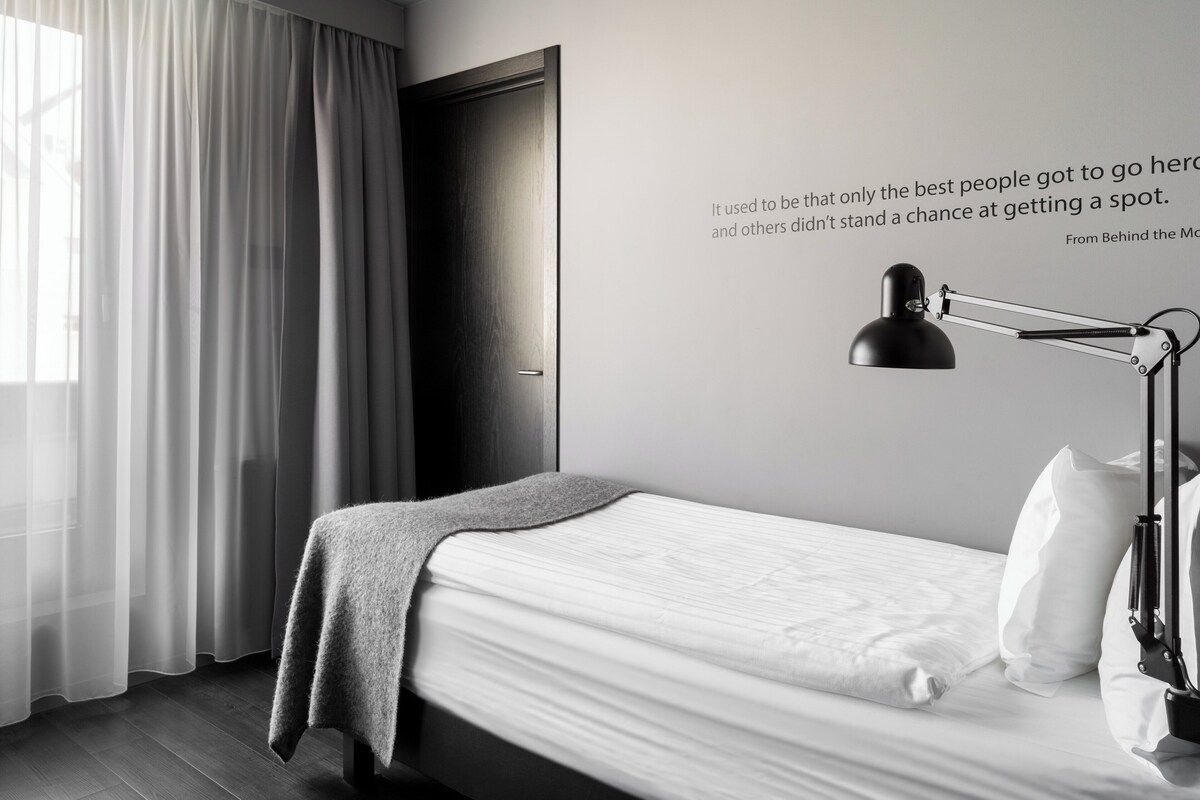Una moderna camera d'albergo con un letto, una lampada da scrivania, una citazione sulla parete e una finestra con tende.