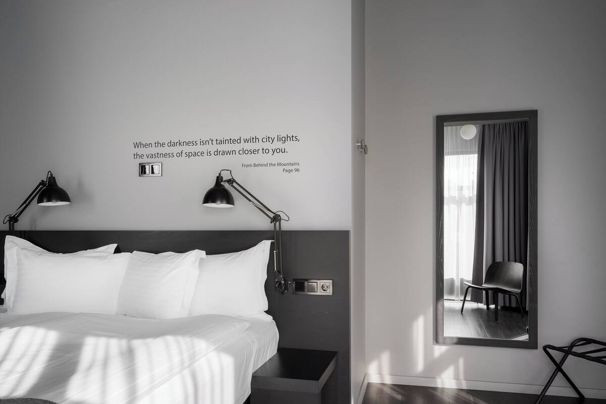 Ein modernes Hotelzimmer mit einem großen Bett, zwei Nachttischlampen, einem Nachttisch, einem Sessel und einem Spiegel. An der Wand ist ein Zitat angebracht.