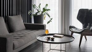 Una sala de estar moderna con un sofá gris, una mesa de centro redonda, un sillón de cuero negro, una planta en maceta y cortinas de tela ligera.