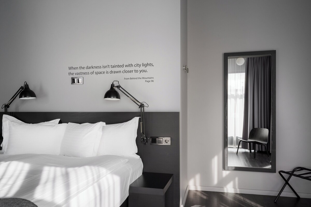 Una moderna camera d'albergo con un letto grande, due lampade da comodino, un comodino, una sedia e uno specchio. Sul muro è esposta una citazione.