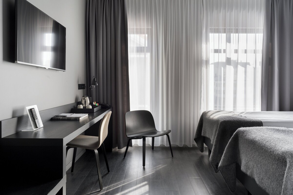 Una camera d'hotel moderna con schermo piatto, una scrivania con sedia, un letto con coperta e ampie finestre con tende leggere.