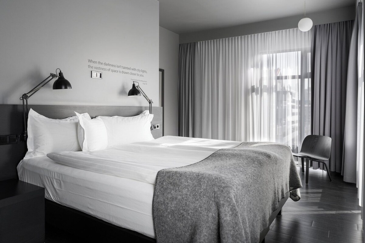 Habitación de hotel moderna con una cama grande, dos lámparas de noche, una silla y vistas a la ciudad a través de cortinas translúcidas.
