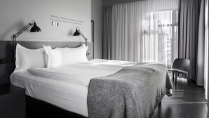 Habitación de hotel moderna con una cama grande, dos lámparas de noche, una silla y vistas a la ciudad a través de cortinas translúcidas.