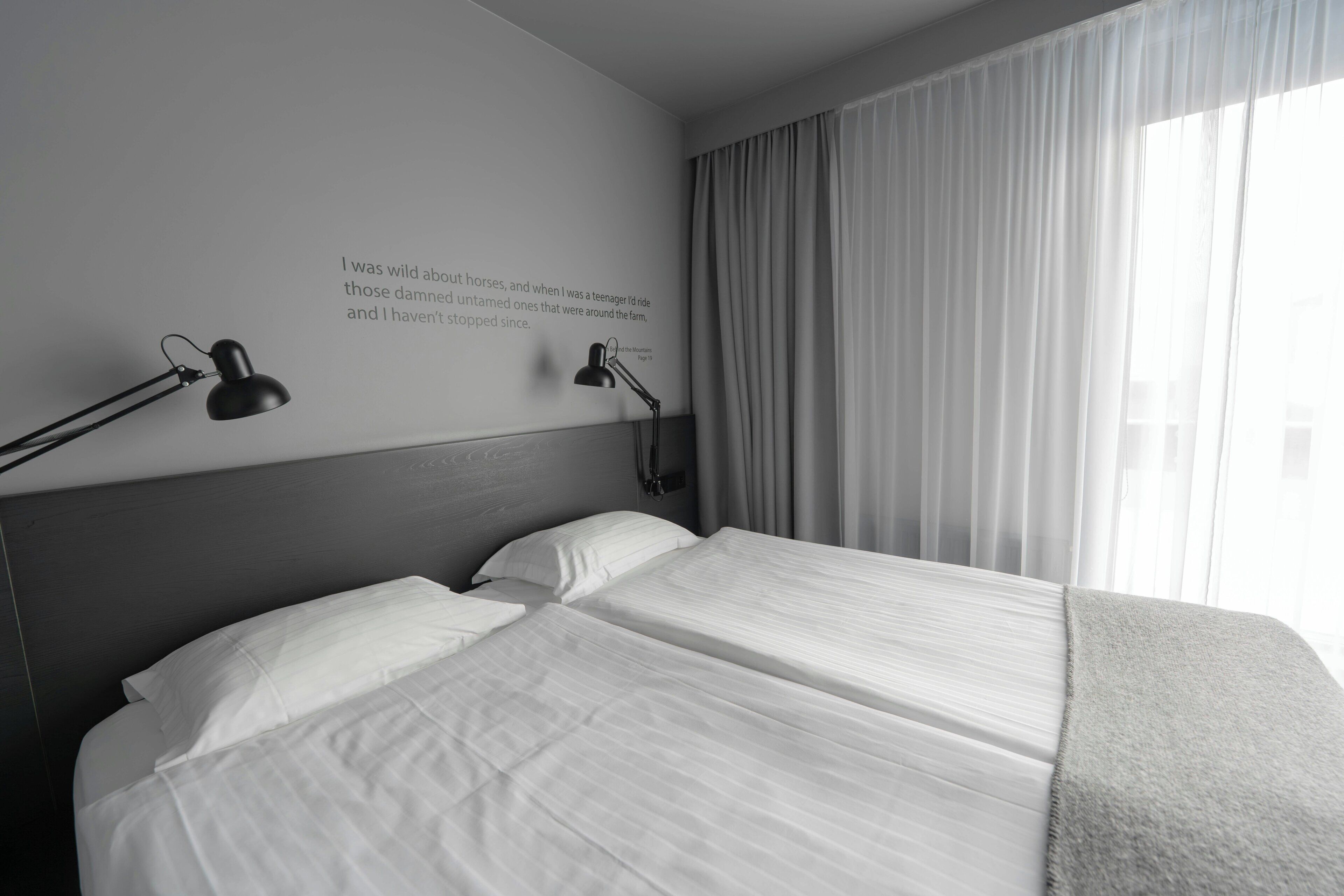 A hotel room with a large bed, two bedside lamps, a wall-mounted reading light, and a window with sheer curtains.