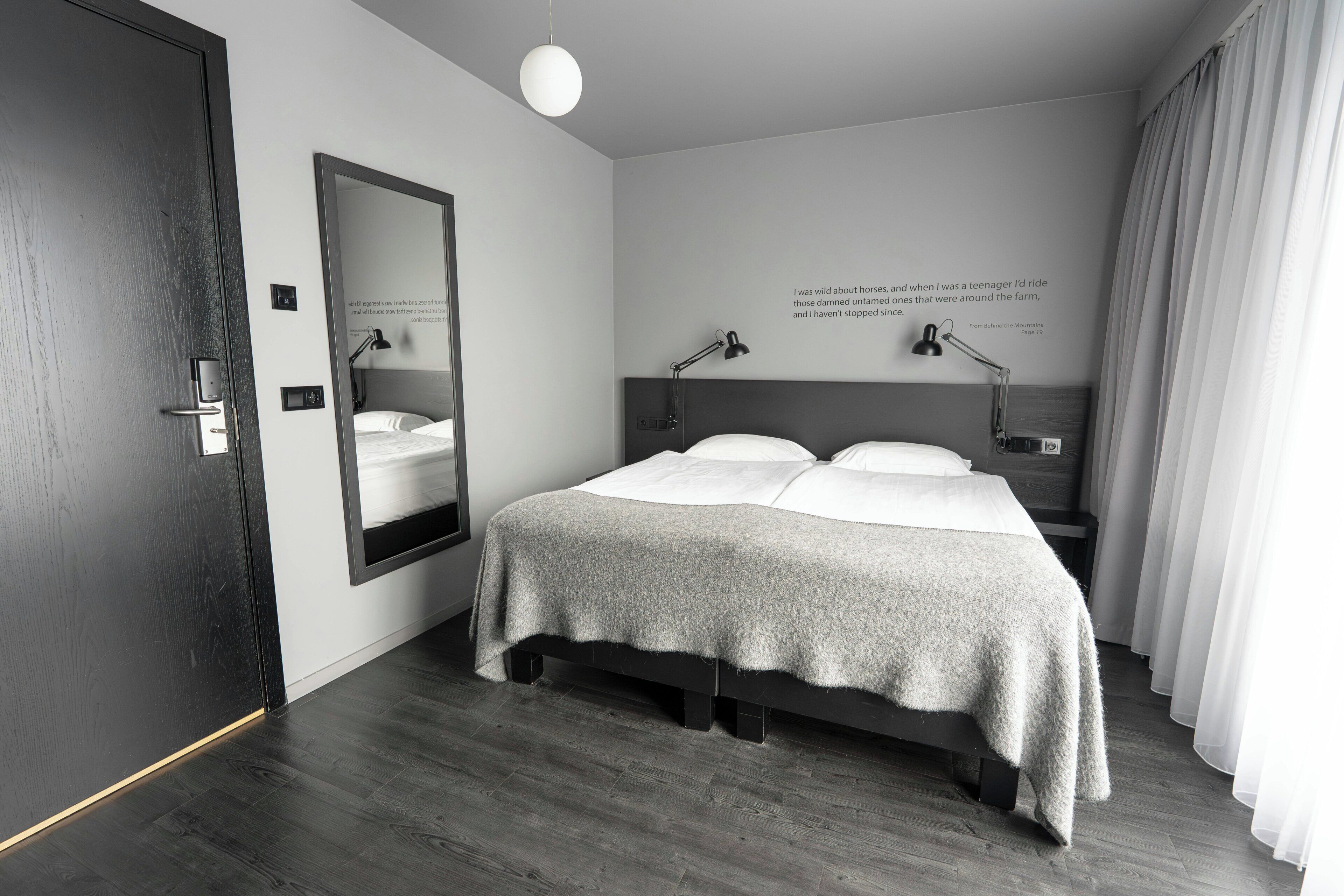 A modern hotel room with a large bed, bedside lamps, a mirror, and a quote on the wall.