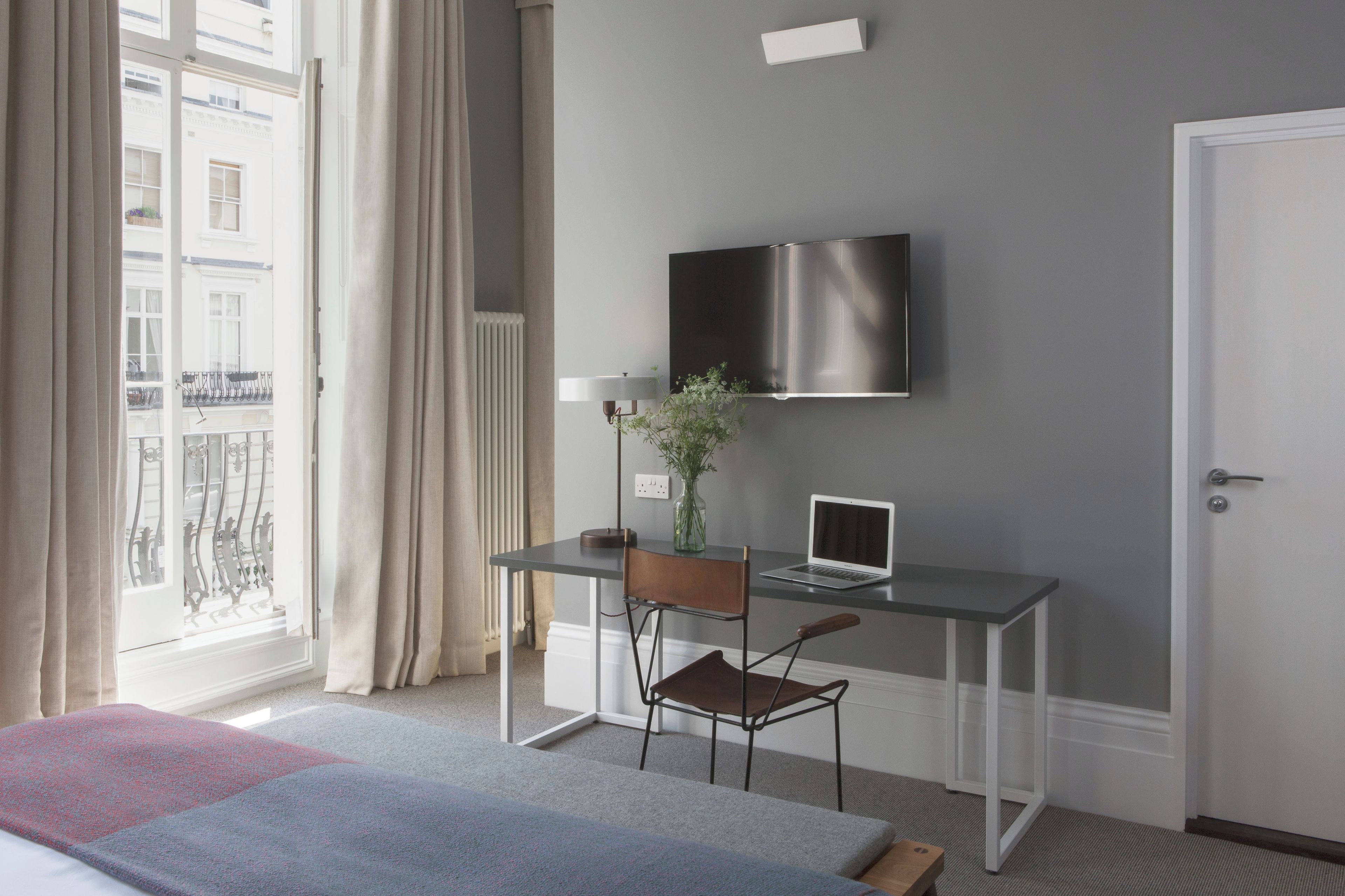 A hotel room with a bed, a desk with a laptop, a chair, a TV mounted on the wall, and a window with curtains.