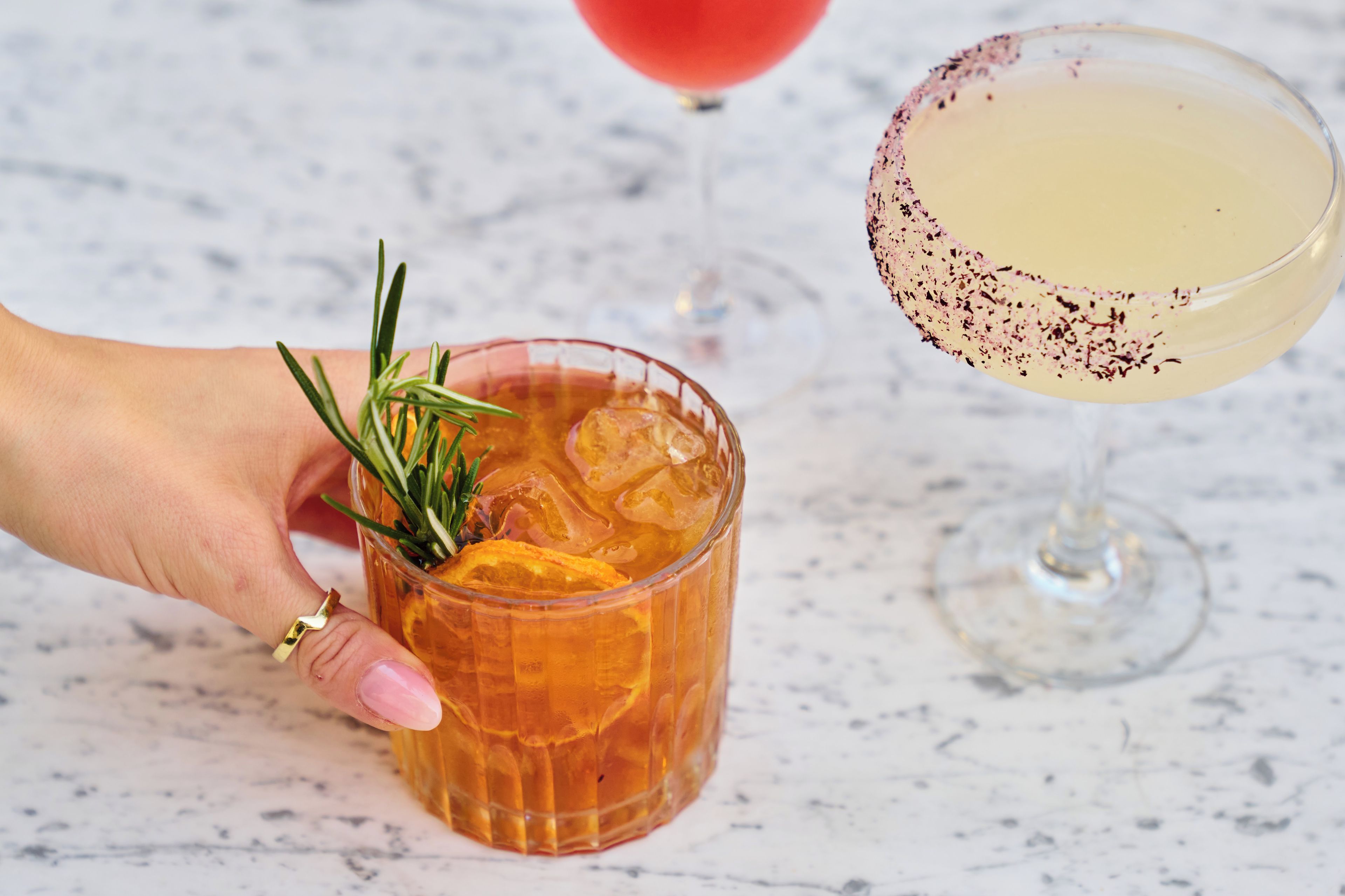 A hand holding a glass with a drink garnished with rosemary and orange slices, and a cocktail glass with a rim dipped in a dark liquid, possibly containing a cocktail.