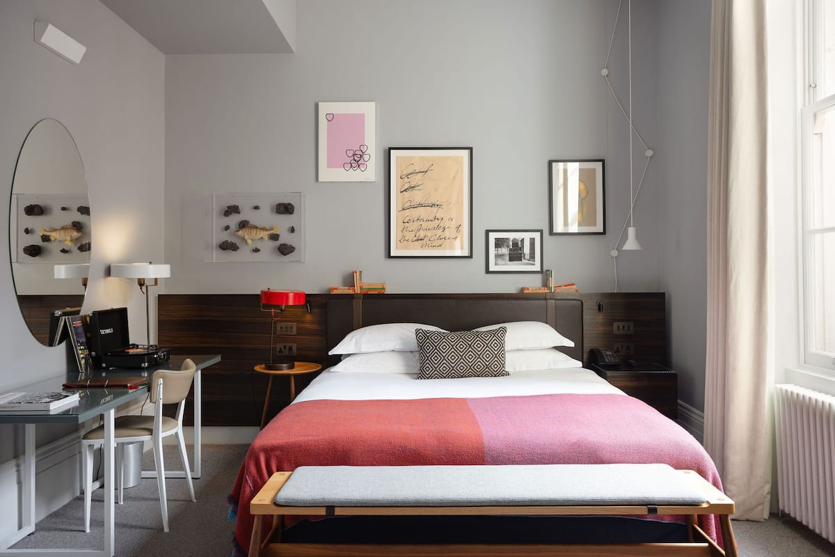A modern bedroom with a bed, bedside tables, a desk, and a chair. There are framed pictures on the wall, a lamp, and a radiator.
