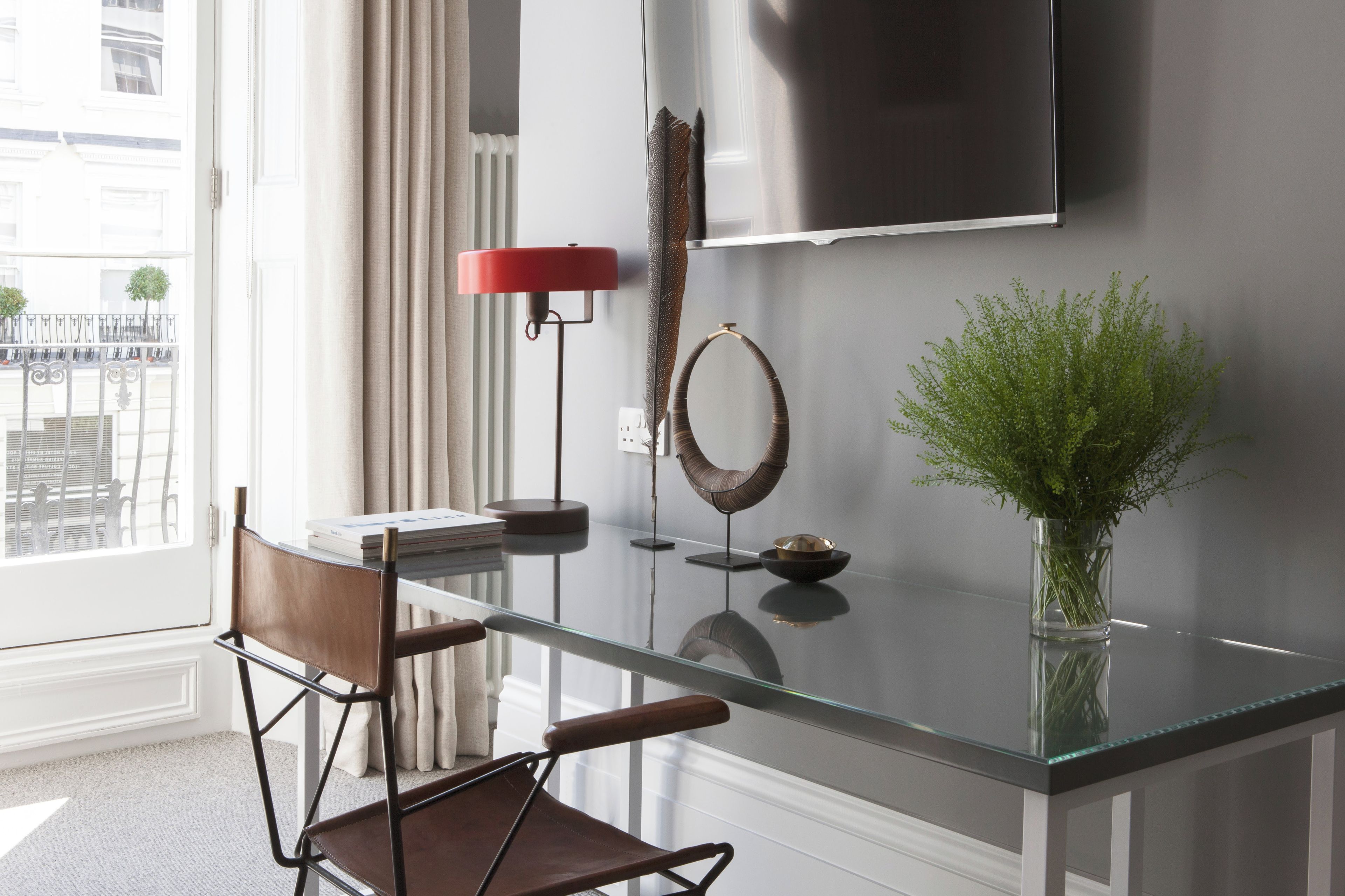 A modern interior with a glass table, two leather chairs, a red floor lamp, a wall-mounted TV, and a vase with green plants.