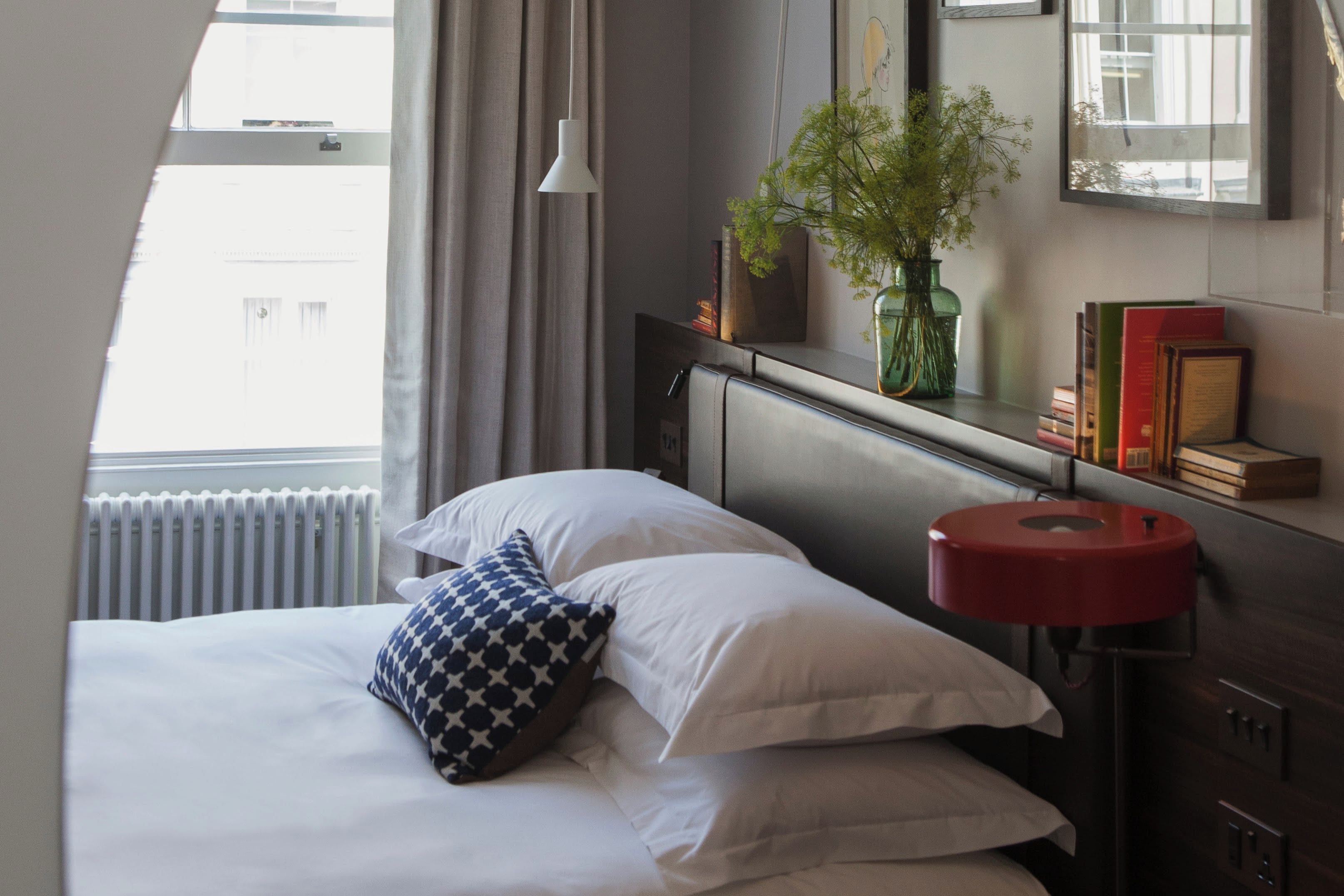 Une chambre d’hôtel avec un lit, une table de chevet sur laquelle se trouve une lampe rouge, un miroir, une fenêtre avec des rideaux et une plante posée sur le rebord de la fenêtre.