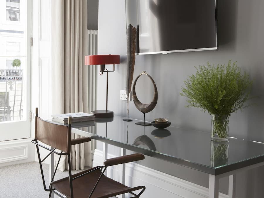 Un salon moderne avec une table basse en verre, un lampadaire rouge, un téléviseur fixé au mur et un vase contenant des plantes vertes.