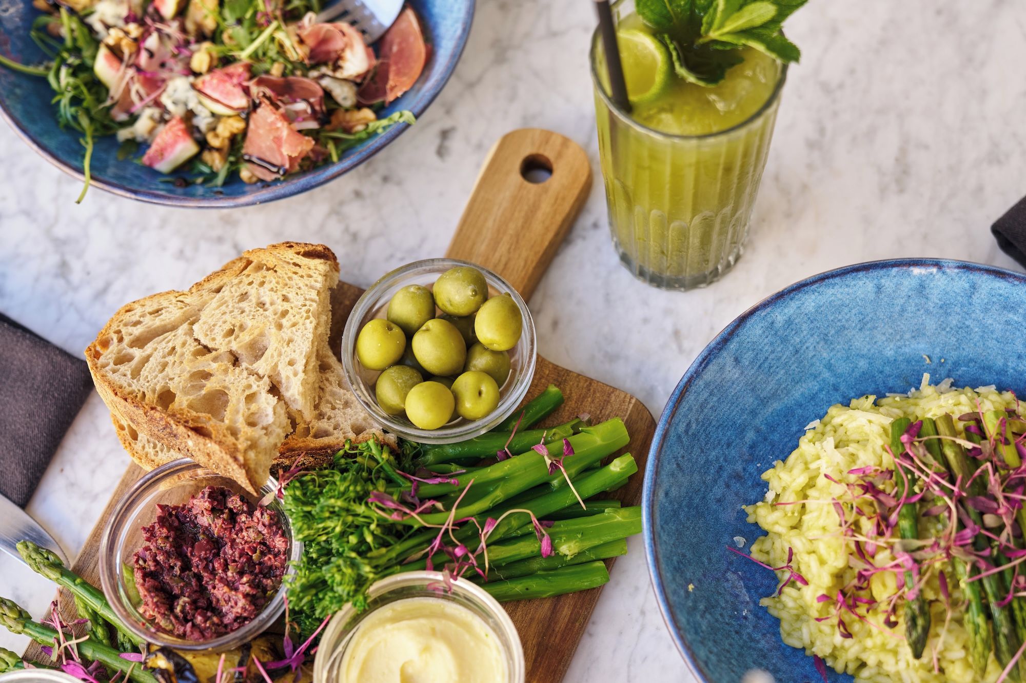 A variety of food items including bread, olives, asparagus, and a green salad on a marble surface.