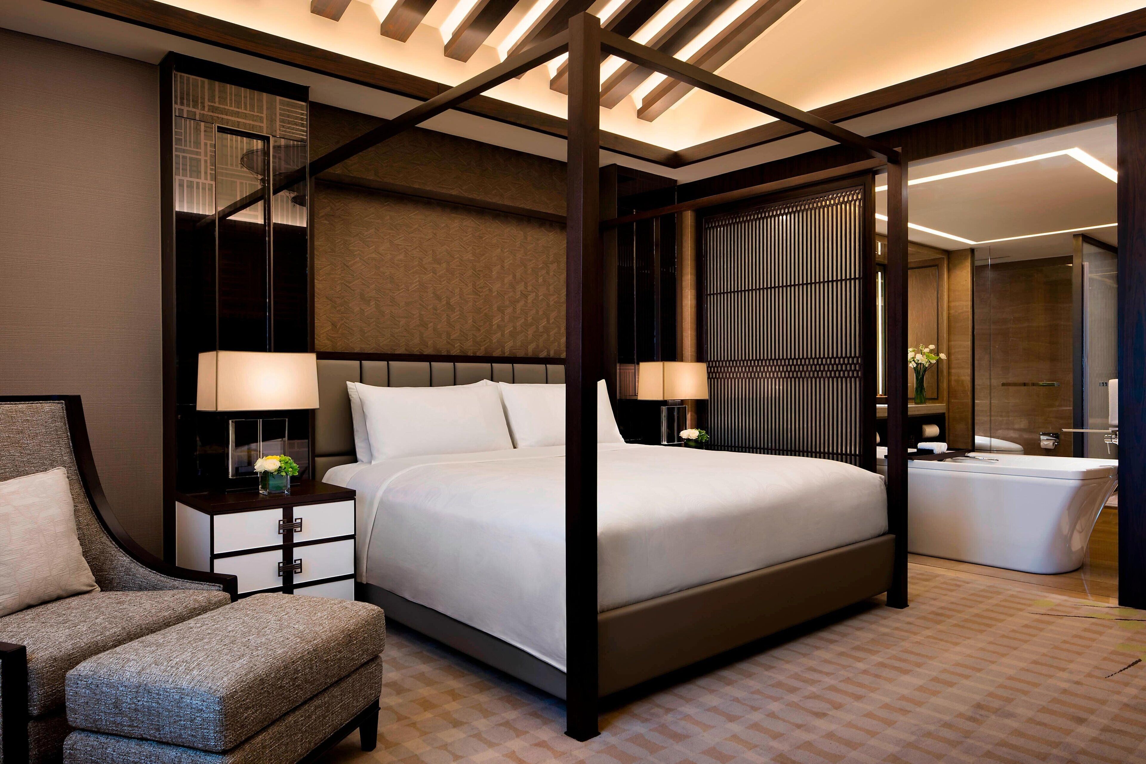 A modern hotel room with a four-poster bed, a sitting area with a chair, a bedside table with a lamp, and a large mirror.