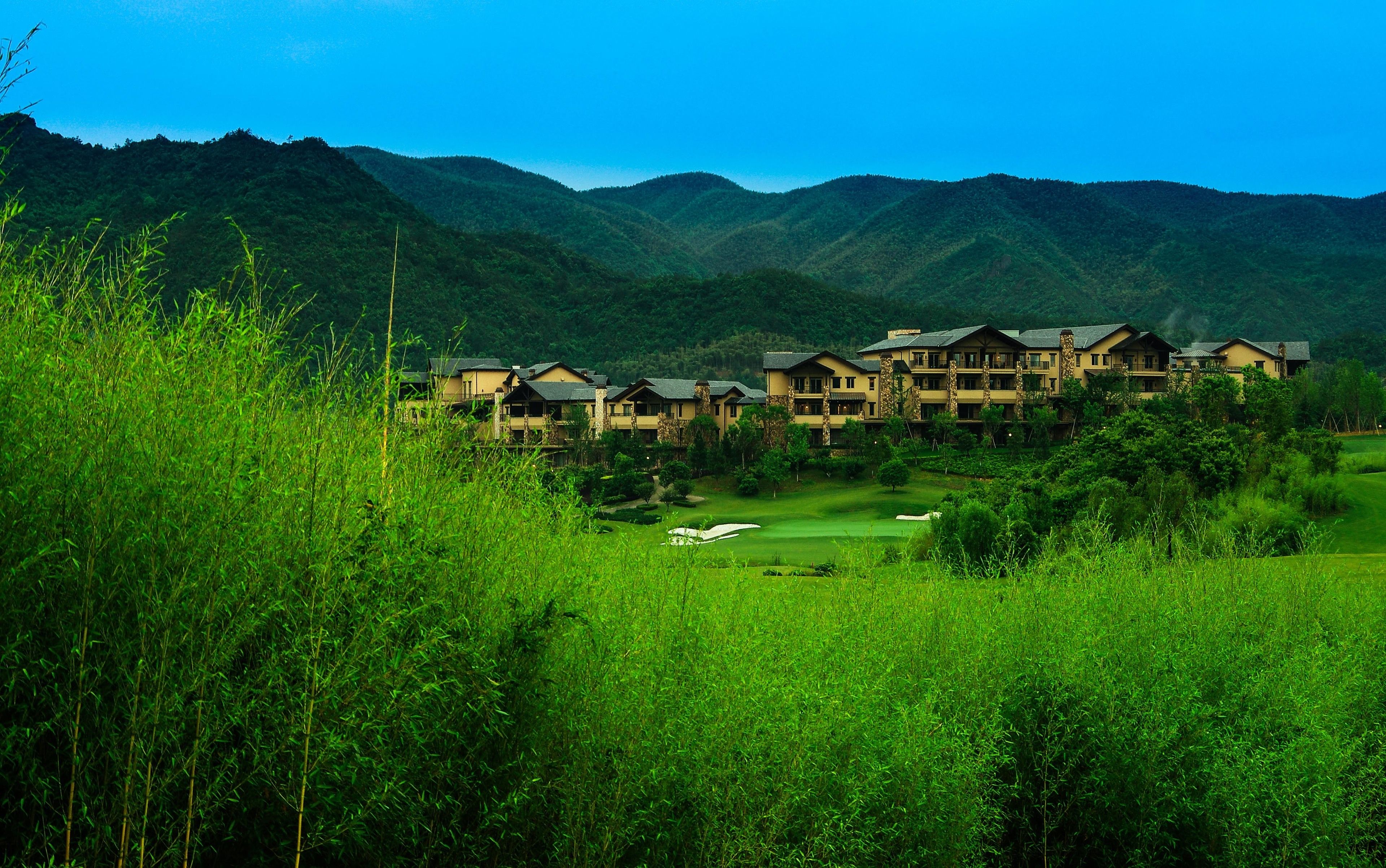 A resort with multiple-story buildings, a golf course, and lush green hills in the background.