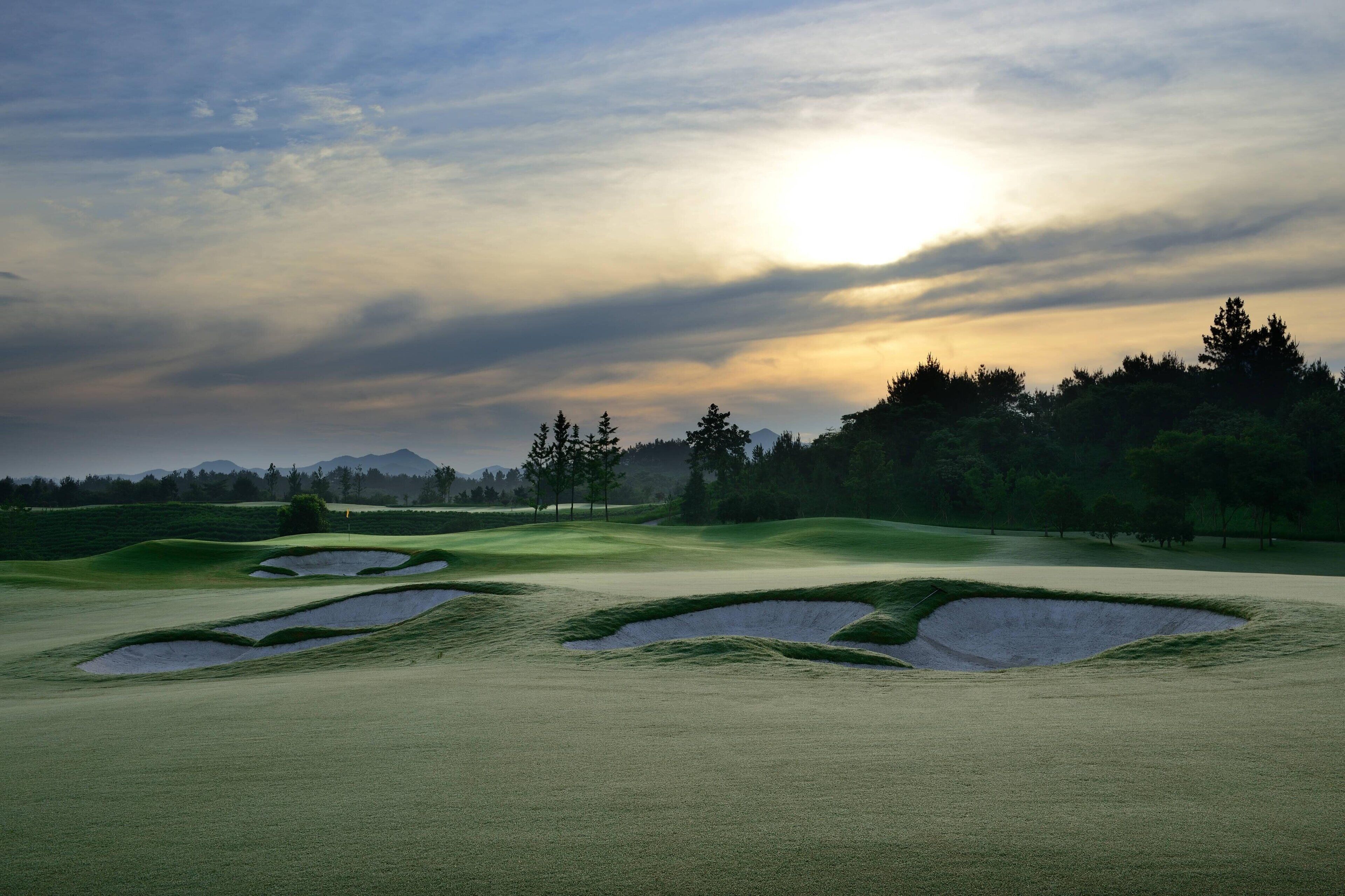 Un parcours de golf avec des bunkers et un mât de drapeau, entouré d’arbres et avec une montagne en arrière-plan.