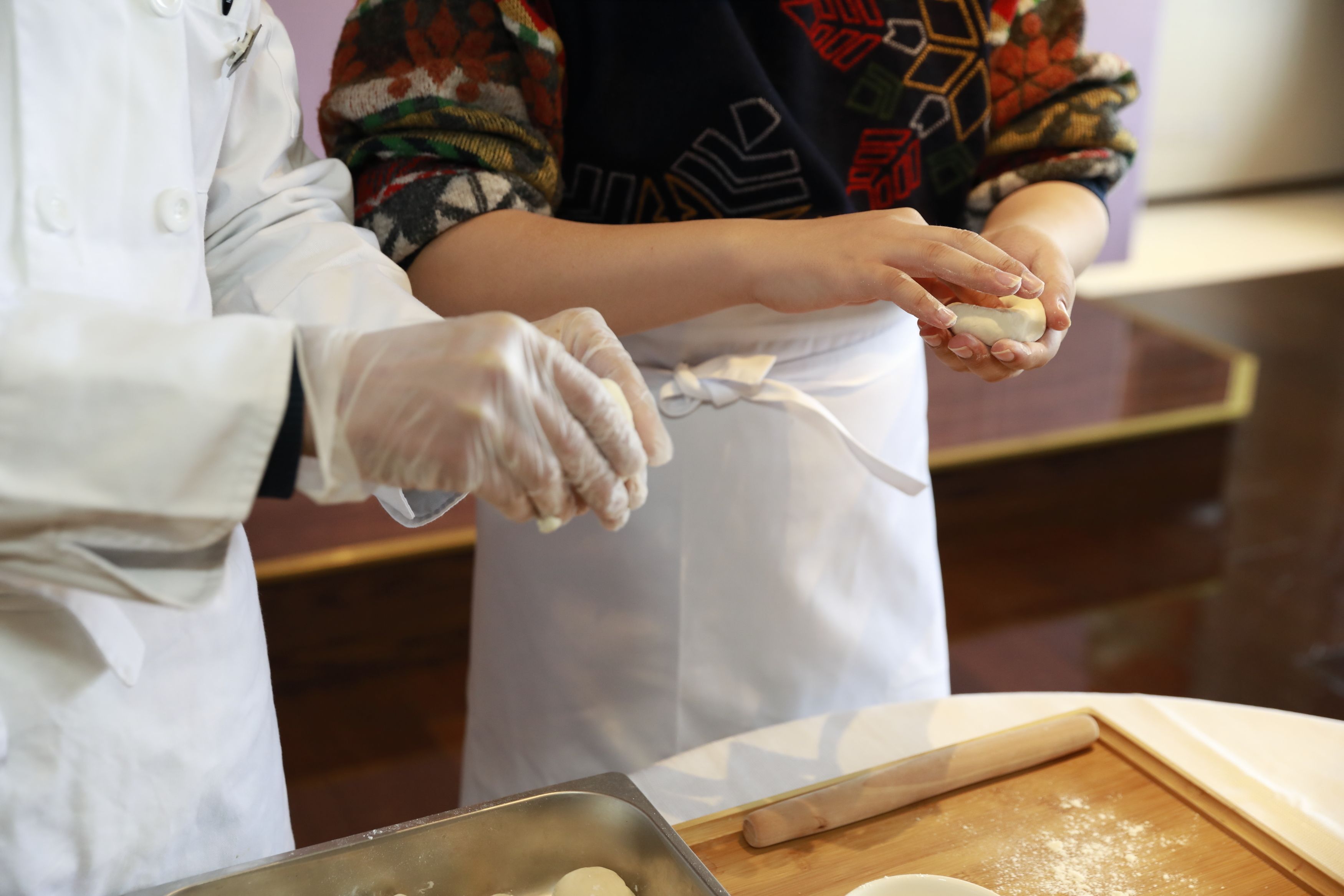 Two individuals in a kitchen preparing food, with a rolling pin and a baking tray on the counter.