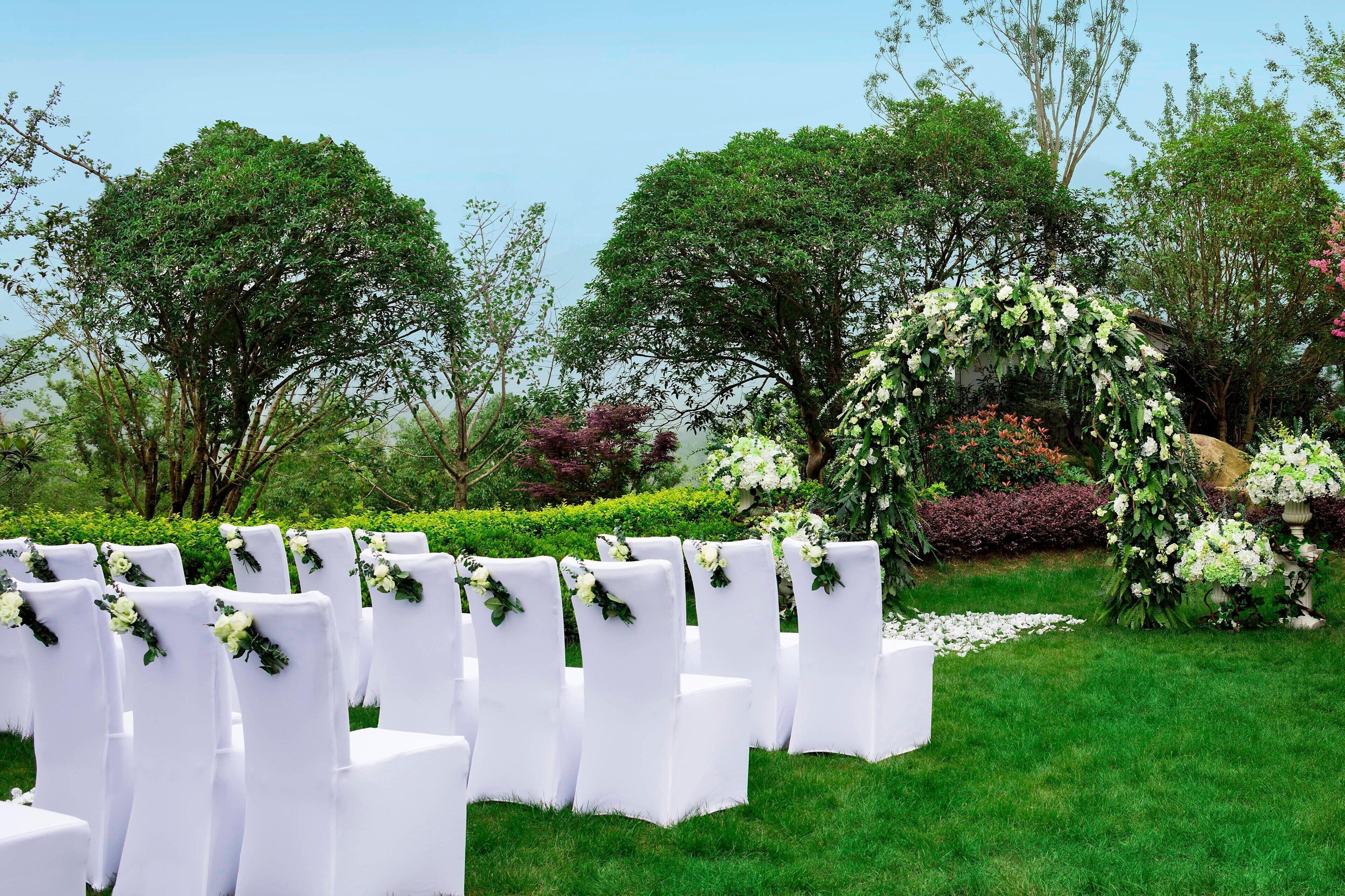 Rows of white chairs with floral decorations, arranged outdoors in a garden setting.
