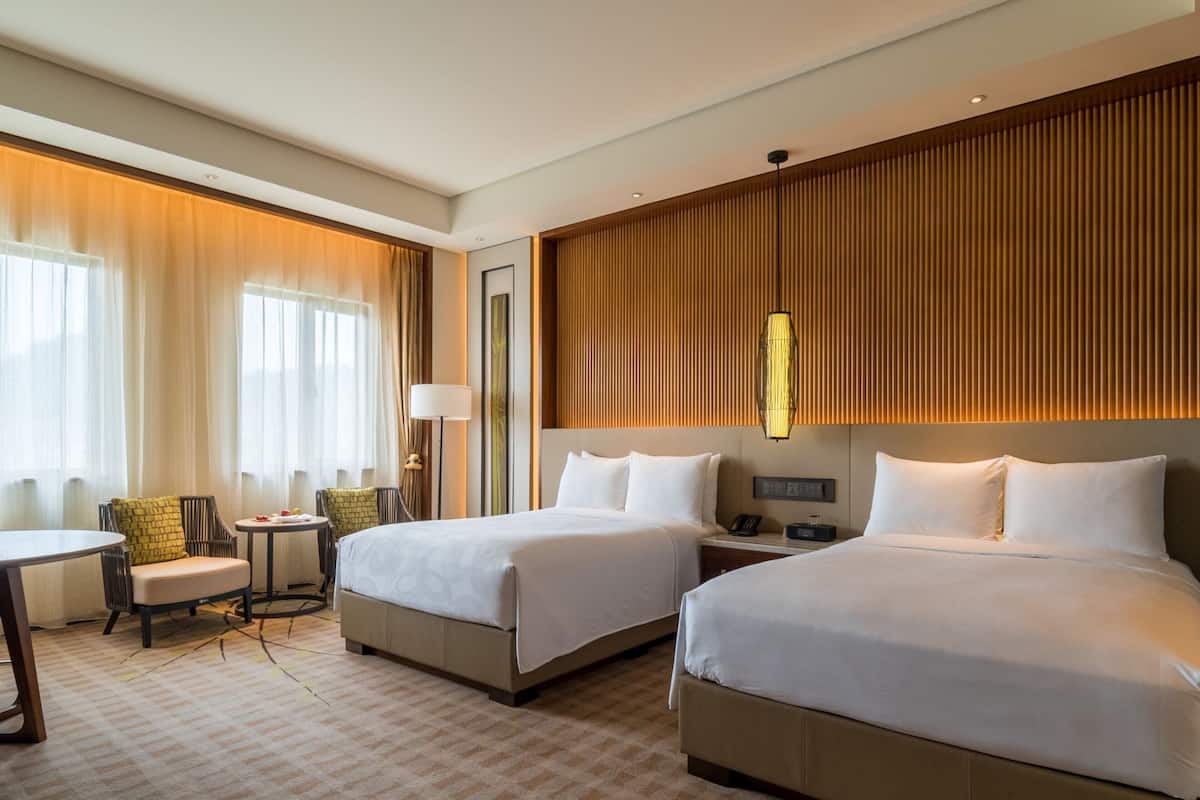 Une chambre d’hôtel avec deux lits, un coin salon comprenant une chaise et une table, et une grande fenêtre avec des rideaux.
