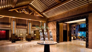 A spacious lobby with a polished marble floor, a central round table with clear glass vases and white flowers, wooden ceiling beams, and a reception desk.