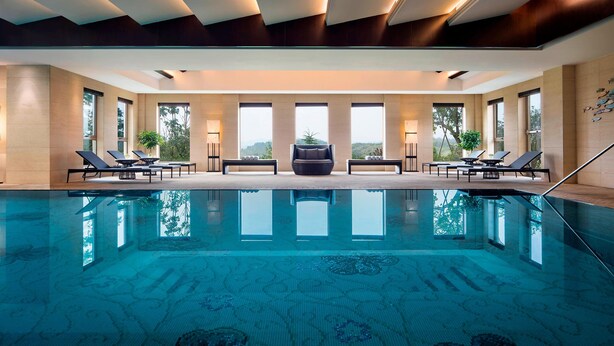 A large indoor swimming pool with lounge chairs and a sofa, surrounded by large windows and a modern ceiling design.