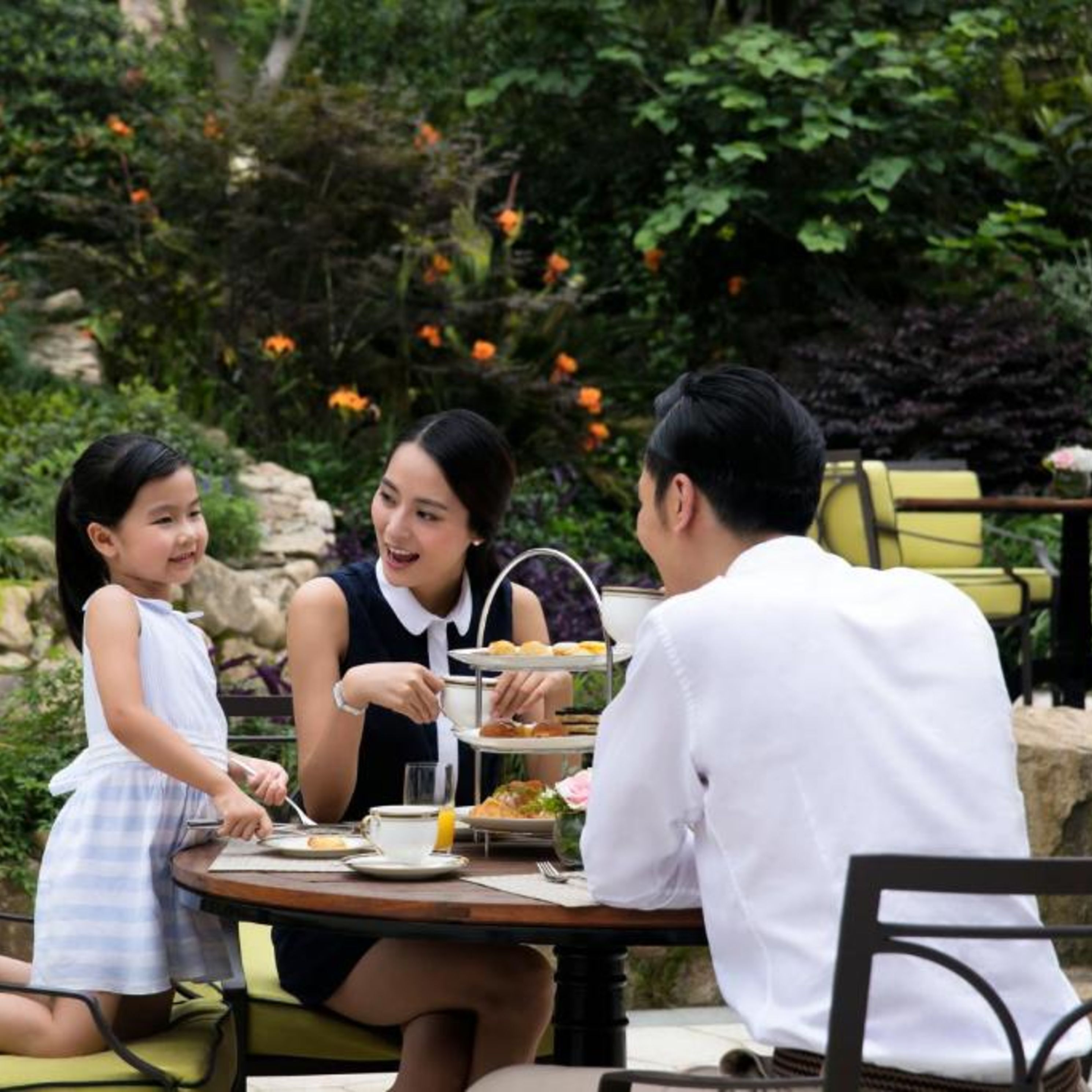 A group of people seated at an outdoor table with a three-tiered tea service, surrounded by greenery and flowers.