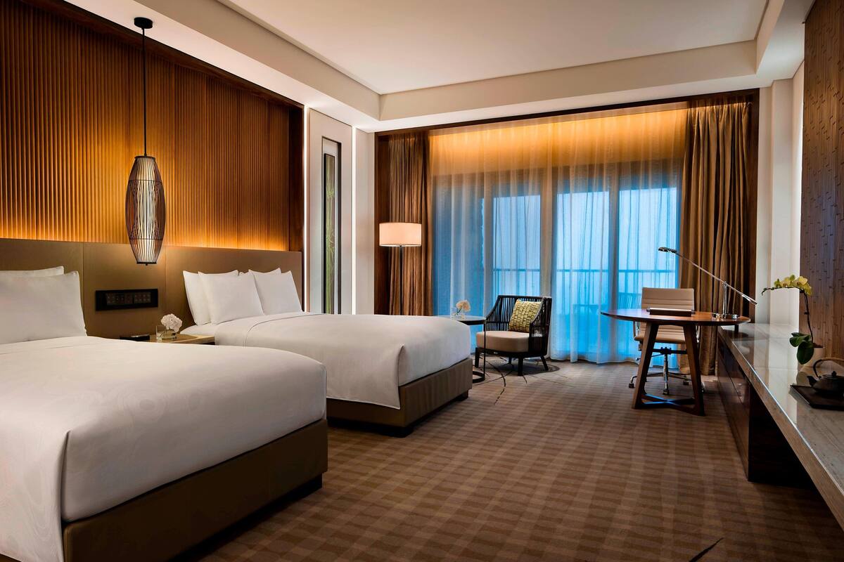Une chambre d’hôtel avec deux lits, un bureau avec une chaise, une lampe et une grande fenêtre avec des rideaux.