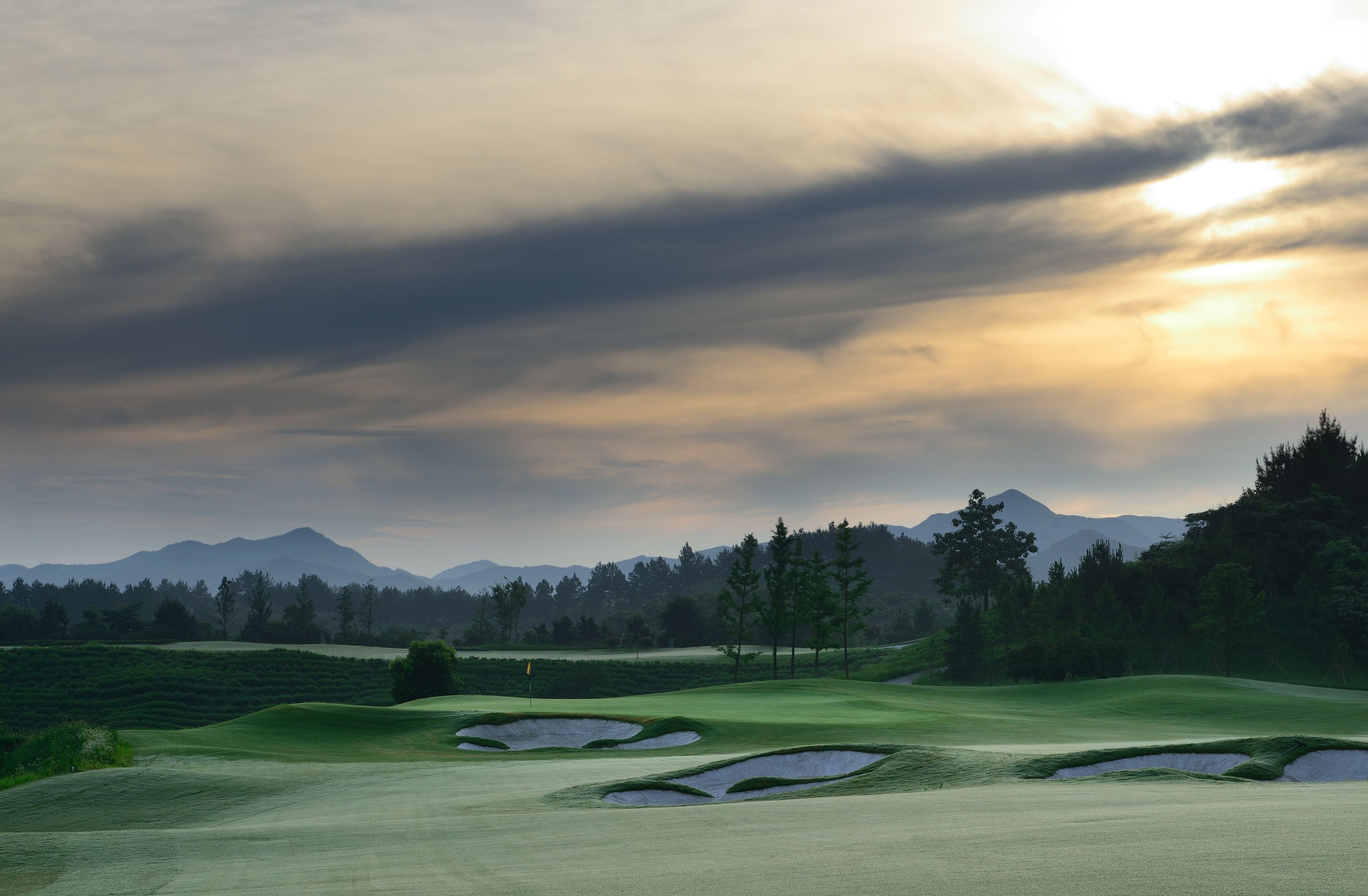 Ein Golfplatz mit Bunkern, Bäumen und in der Ferne Bergen unter einem bewölkten Himmel.