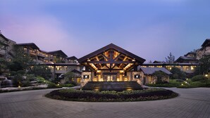 A hotel with a central building featuring a stone facade and a wooden upper structure, surrounded by landscaped gardens and a paved walkway.